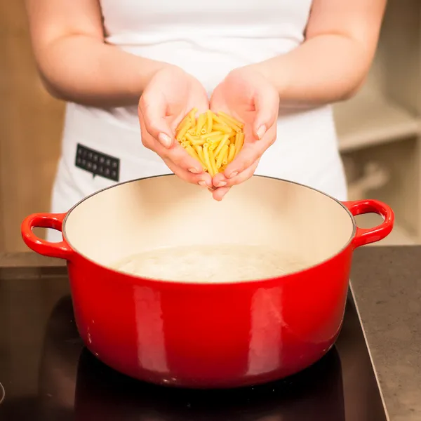 Person holding dry yellow pasta over a red pot filled with boiling water on a stovetop.