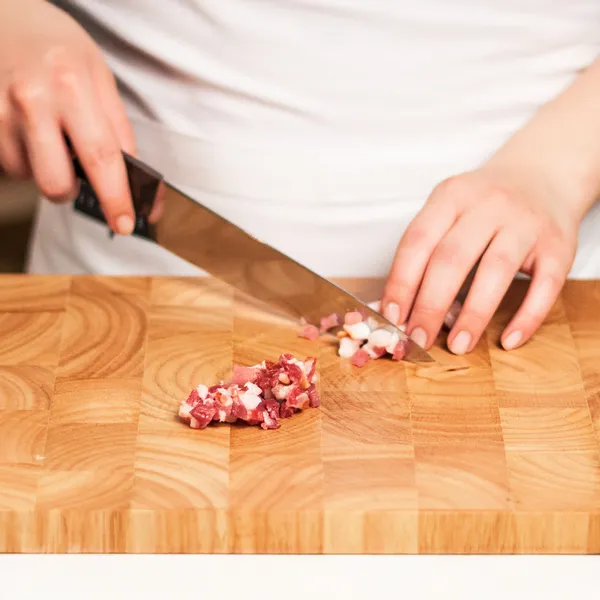 Person finely chopping raw meat on a large wooden cutting board using a kitchen knife.