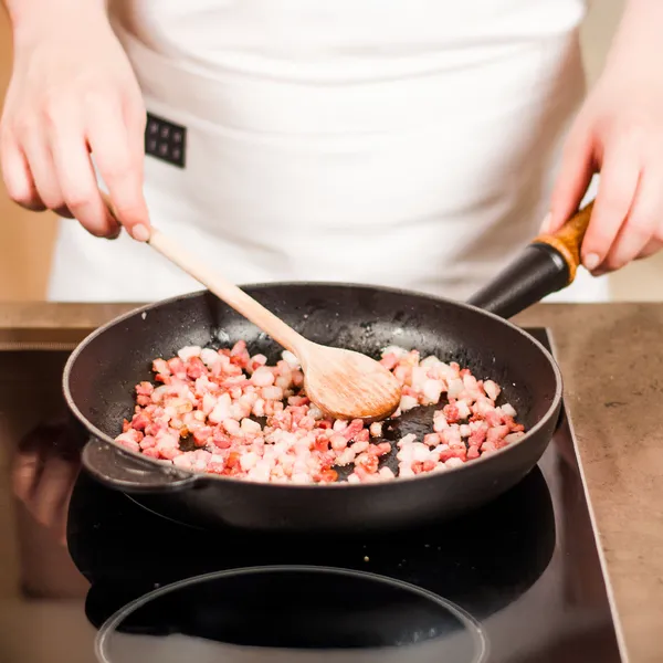 Person stirring diced bacon in a black frying pan on an electric stove with a wooden spoon.