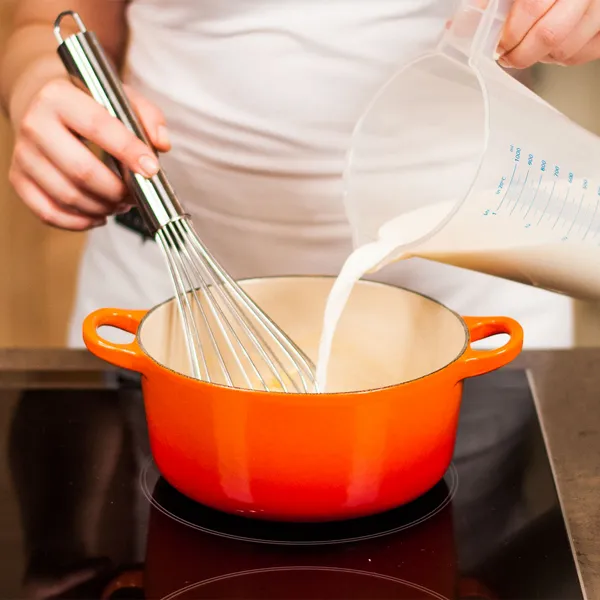 Person pouring milk into an orange pot on a stove while holding a whisk, preparing a recipe.