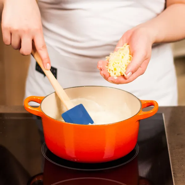 Person adding shredded cheese to a creamy mixture in an orange pot on a stovetop, stirring with a blue spatula.