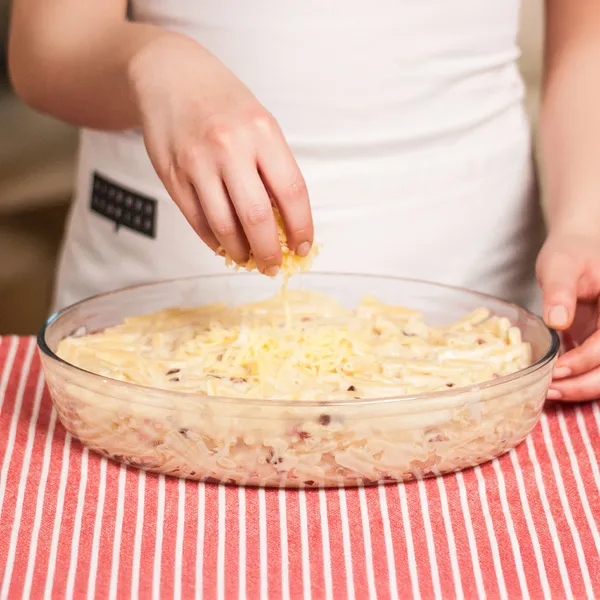 Hand sprinkling grated cheese over a creamy pasta dish in a clear glass baking dish on a red-striped cloth.