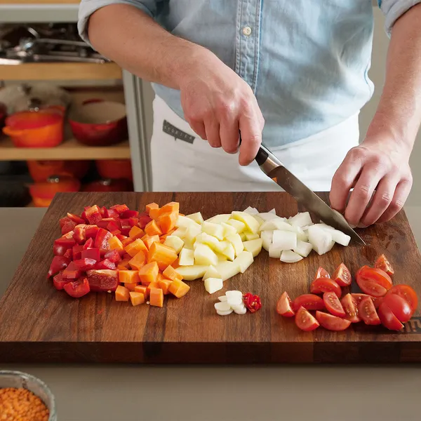 Person chopping various fresh vegetables including tomatoes, onions, carrots, and garlic on a wooden cutting board in a kitchen.
