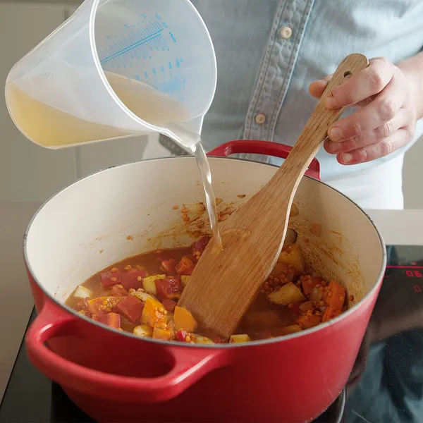 Hands pouring clear broth from a plastic measuring jug into a red enameled pot of chopped vegetables while stirring with a wooden spoon