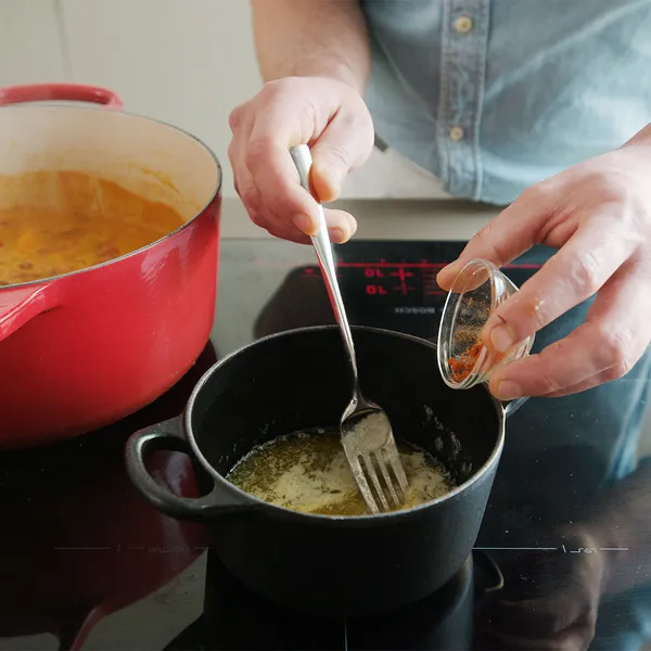 Person’s hands stirring melted butter with a fork in a small black saucepan while sprinkling red spice from a tiny jar