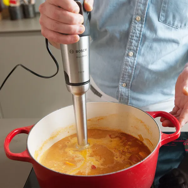 Hands using a stainless immersion blender to puree orange soup in a red enameled pot on an induction cooktop