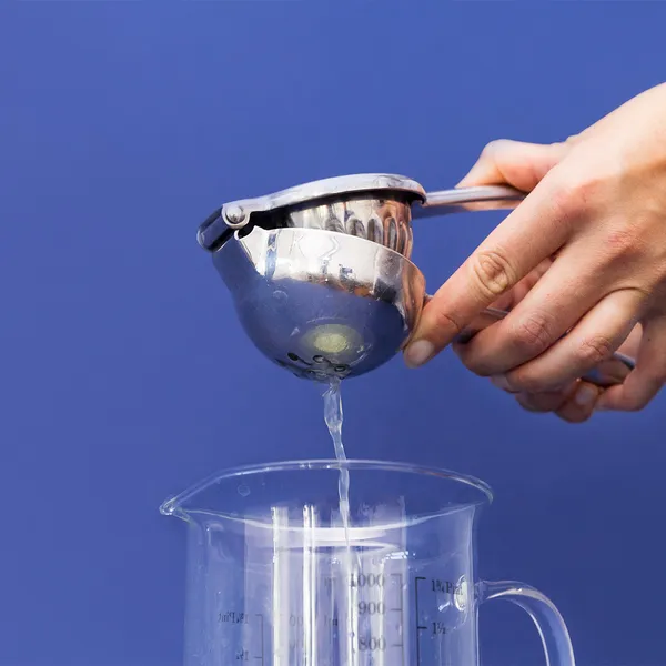 Hand squeezing lemon juice through a stainless steel citrus press into a clear measuring jug against a blue background.