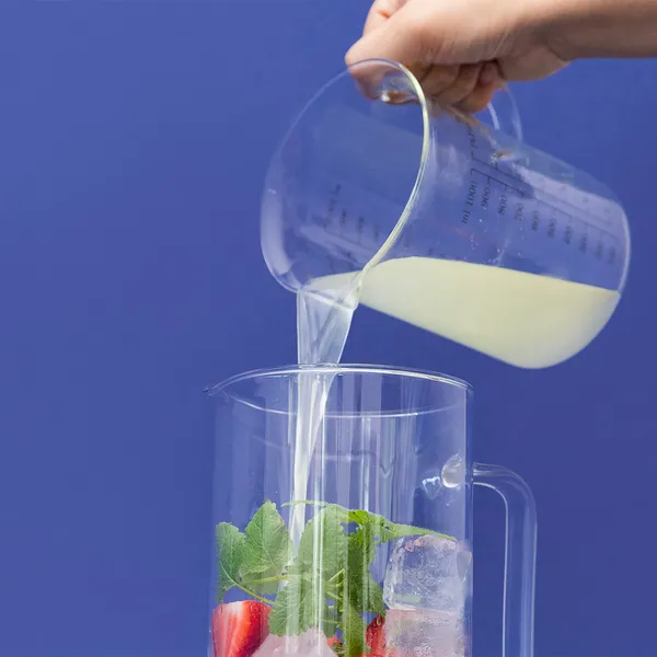 Hand pouring milk from a measuring jug into a blender containing fresh fruit and leafy greens against a blue background.