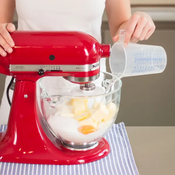 Person pouring liquid from a measuring cup into a red stand mixer bowl containing eggs, sugar, and butter