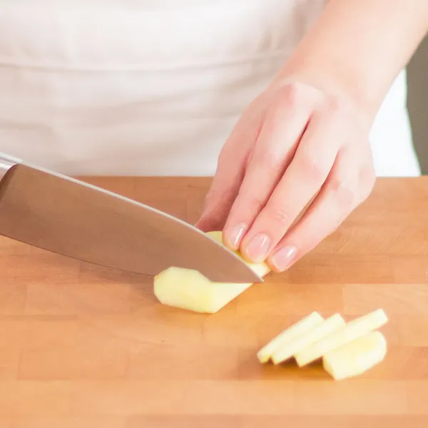 Hand slicing a peeled potato into thin pieces on a wooden cutting board with a kitchen knife