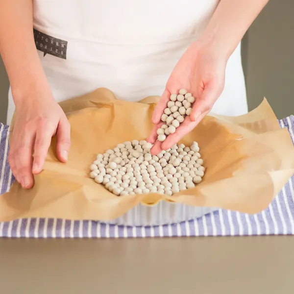 Hands spreading ceramic baking beans on parchment paper in a baking dish on a striped cloth