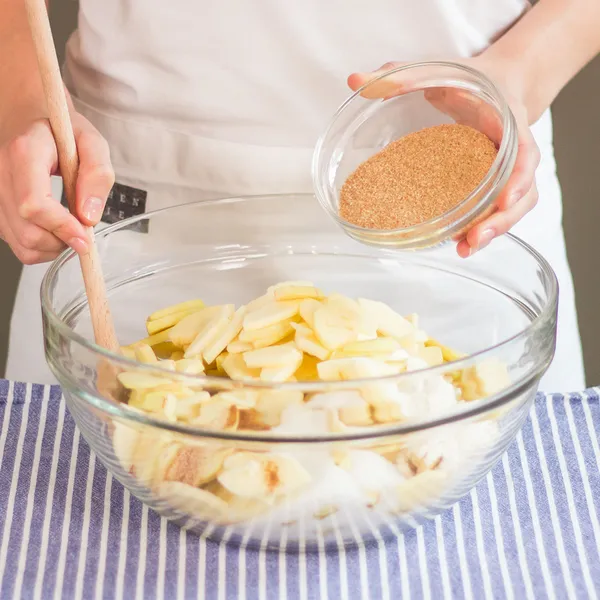 Person stirring sliced apples in a large glass bowl while holding a small glass bowl of brown sugar above it