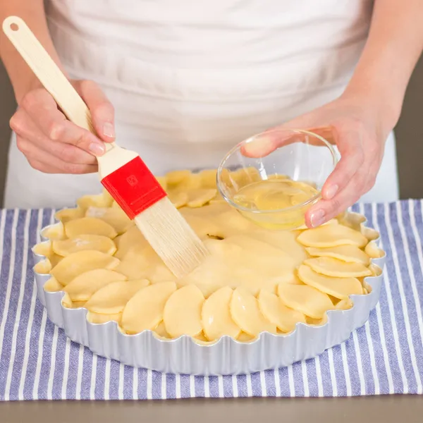 Person brushing liquid onto a round apple tart with a red-handled pastry brush