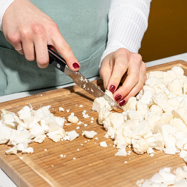 Close-up of hands with red nail polish chopping cauliflower into florets on a wooden cutting board with a knife.
