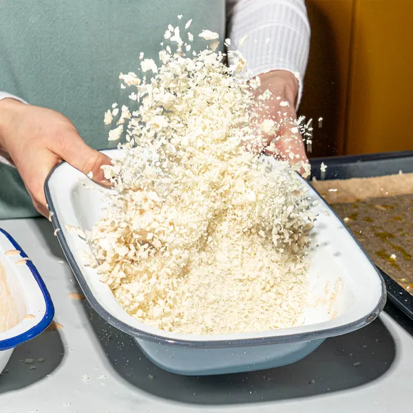 Hands shaking a tray of panko breadcrumbs over a table with a batter bowl and baking sheet nearby