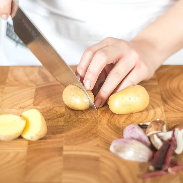 Hands cutting small potatoes with a knife on a wooden board; shallots and mushrooms beside them.