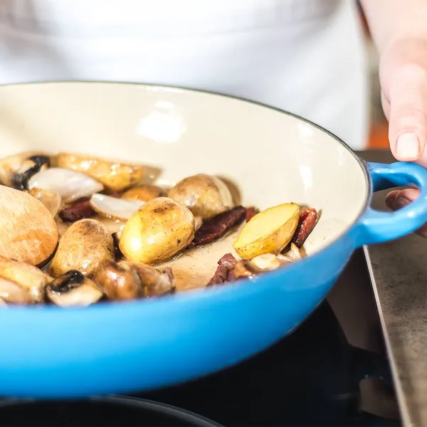Blue casserole with small potatoes, mushrooms and sautéed shallots; one hand holds the handle and the other stirs with a wooden spoon.