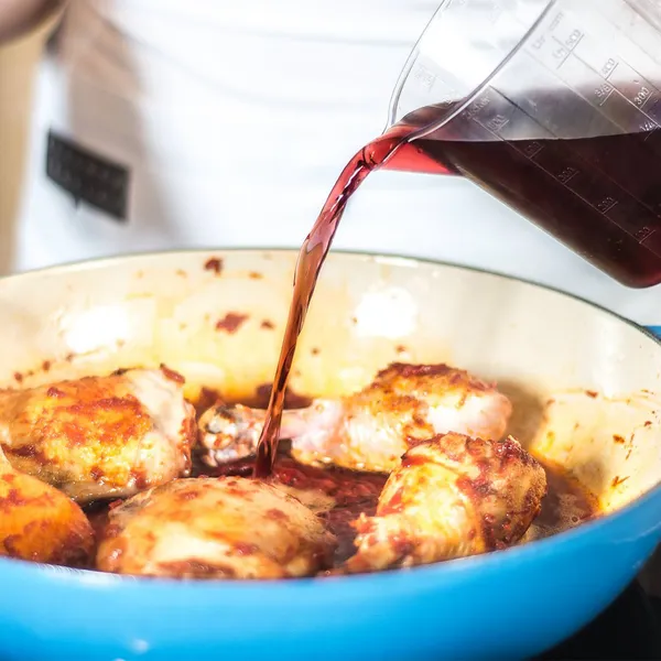 Measuring jug pouring red wine into a blue casserole with browned chicken thighs and a wooden spoon.