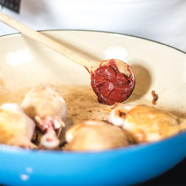 Close-up of a blue enameled casserole with browned pieces of chicken; a hand holds a wooden spoon adding a spoonful of tomato paste to the bottom of the pot.