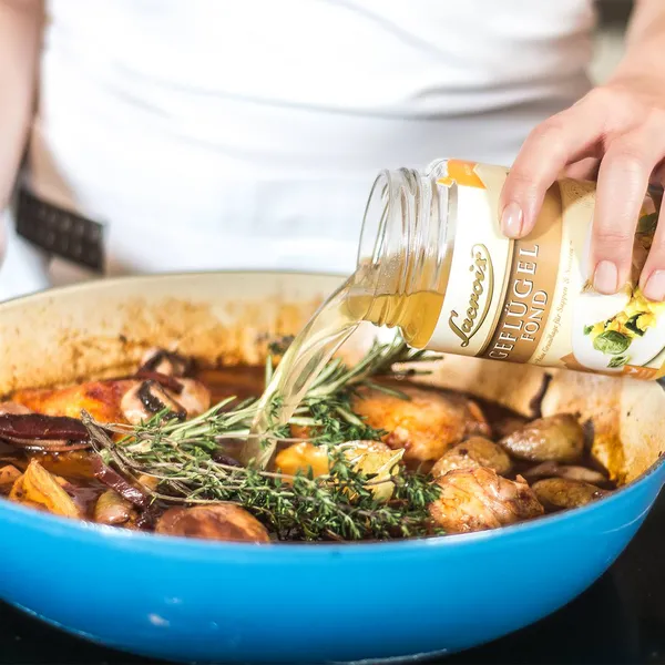Close-up of hands pouring chicken stock from a jar into a blue enameled casserole with browned chicken thighs, potatoes, mushrooms and sprigs of thyme.