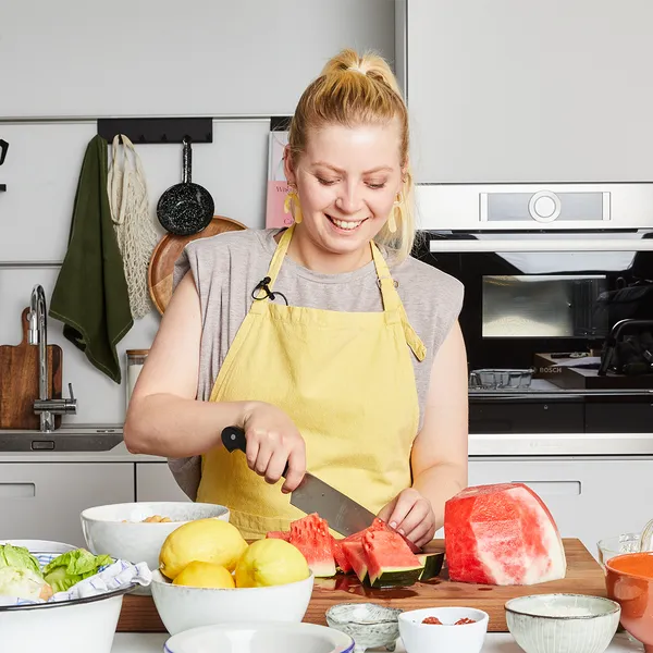 Person wearing a yellow apron cutting watermelon on a kitchen counter with various bowls and fruits around