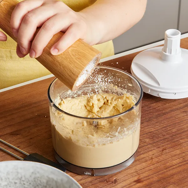 Hands grinding seasoning into a glass container with dough inside on a wooden surface