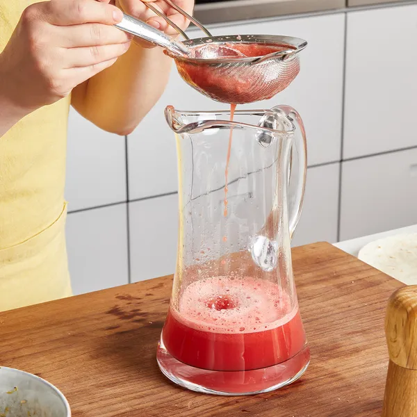 Person straining red liquid through a metal sieve into a glass pitcher on a wooden countertop