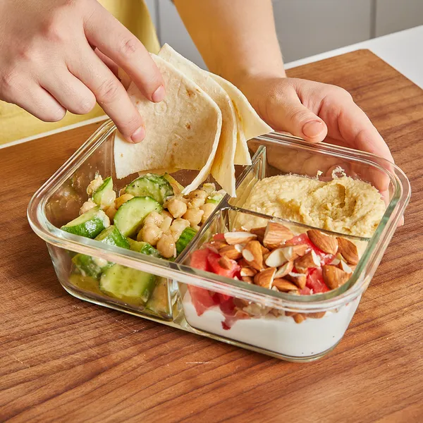 Person dipping flatbread into a divided glass container with cucumber chickpea salad, hummus, and yogurt topped with almonds and fruit.