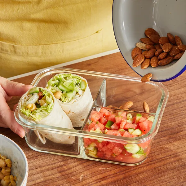 Glass divided container with two wrap halves filled with lettuce and chickpeas and a section of diced cucumber and tomato with almonds being poured from a white bowl