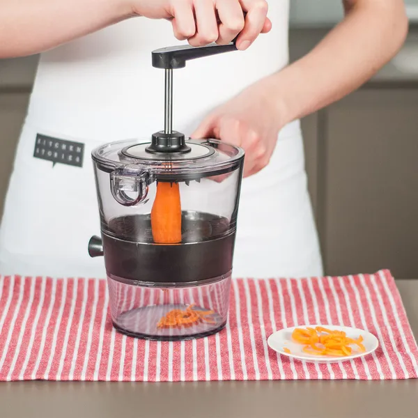 Person using a manual food processor to grate a carrot on a red-and-white striped cloth with a small plate of grated carrot nearby.