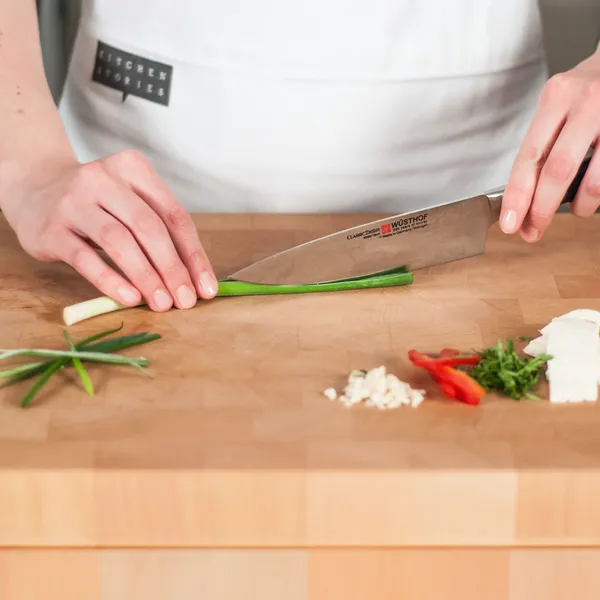 Hands holding a knife slicing a green onion on a wooden cutting board with small piles of chopped herbs and vegetables nearby.