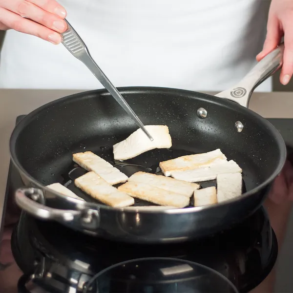 Hands holding metal tongs cooking rectangular pieces of food in a frying pan on a stovetop.