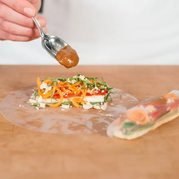 Hand holding a spoon adding sauce over a rice paper sheet topped with sliced vegetables and herbs on a wooden surface.