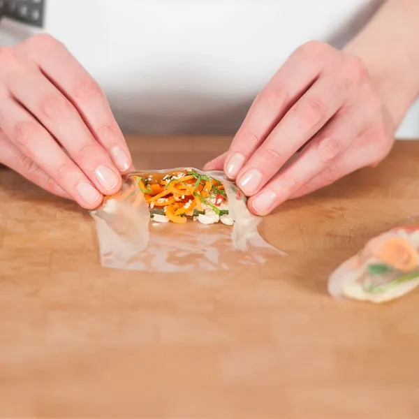 Hands folding a translucent rice paper wrapper around a filling of shredded vegetables on a wooden surface.
