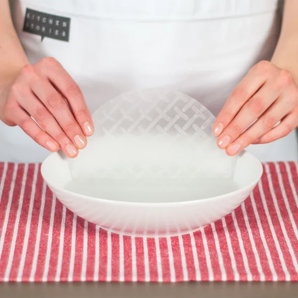 Hands holding a translucent rice paper sheet over a white bowl on a red-and-white striped cloth.