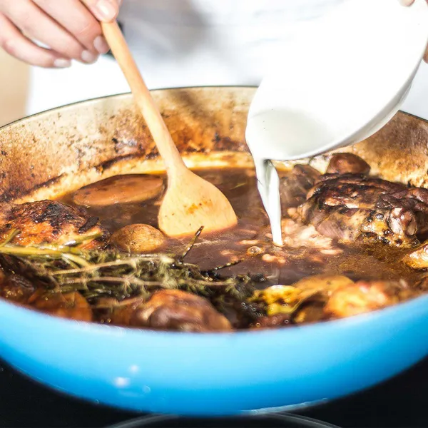 Close-up of hands pouring cream into a blue enameled casserole with browned chicken thighs, potatoes, mushrooms and sprigs of thyme; a wooden spoon is in the stew.