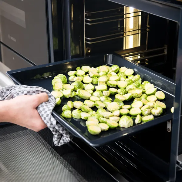 Hand holding a baking tray with halved Brussels sprouts being placed into an open oven