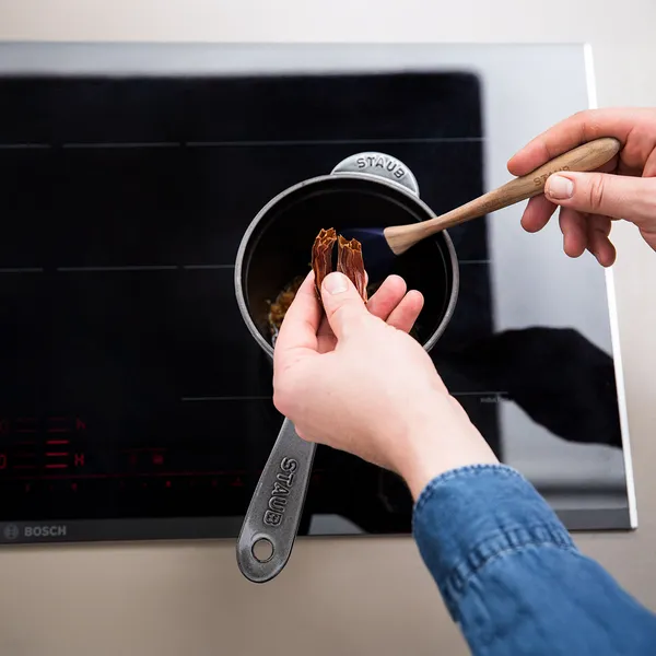 Hands holding dried bay leaves over a small pot on a stovetop with a wooden spoon inside the pot
