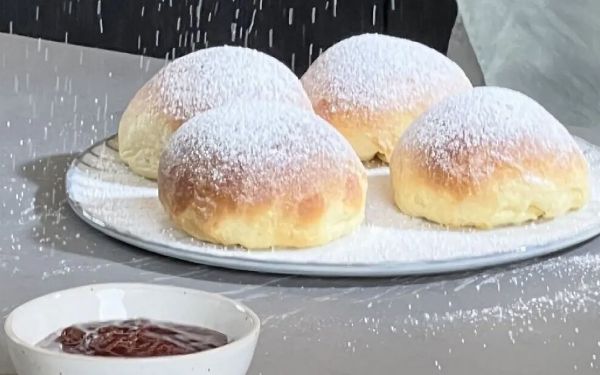 Vier mit Puderzucker best&auml;ubte Krapfen auf einem wei&szlig;en Teller neben zwei Schalen mit dunkler und roter Fruchtf&uuml;llung