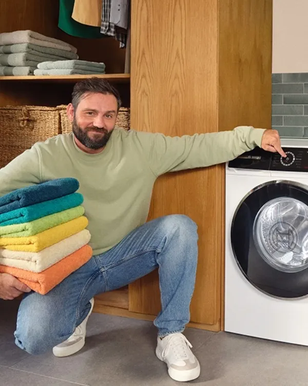 Man holding colorful folded towels, squatting next to a washing machine and dryer in a modern laundry room.