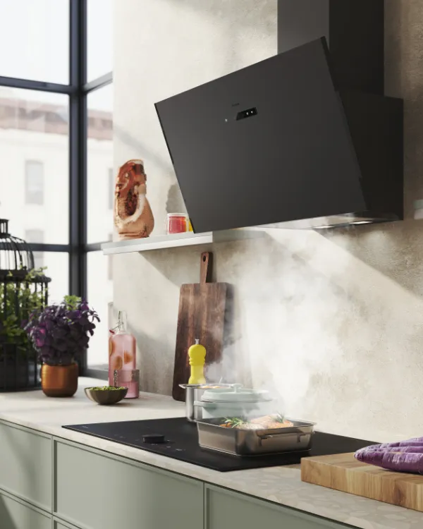Modern kitchen with green cabinets, black angled range hood, wooden utensils, potted plants, and food on the countertop near large windows.