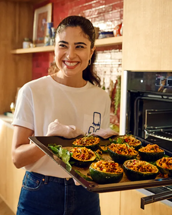 Smiling woman holds a tray of stuffed squash, ready to place them in an open oven in a modern kitchen.