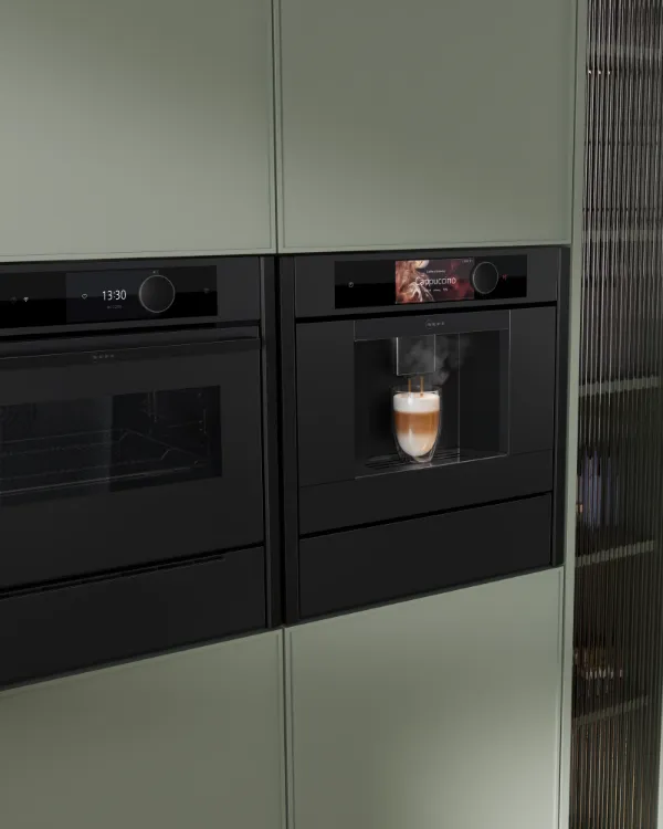 Built-in black coffee machine brewing a cappuccino next to a black oven in a modern kitchen with green cabinets.