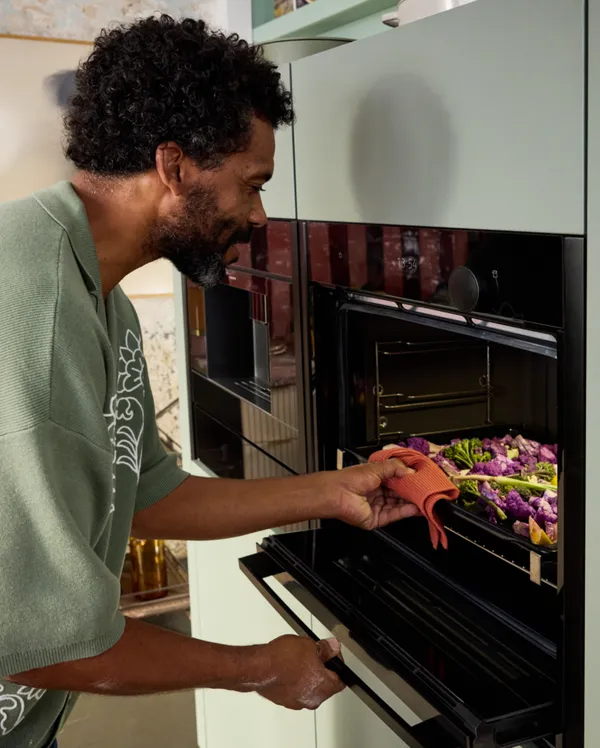 Man with curly hair and a beard leans into a built-in oven, using a red dish towel to pull out a tray. The tray holds purple cauliflower and green vegetables.