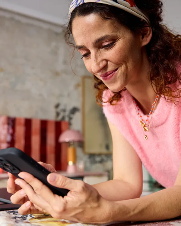 Smiling woman in a pink sleeveless sweater leaning on a kitchen counter, looking at her phone.