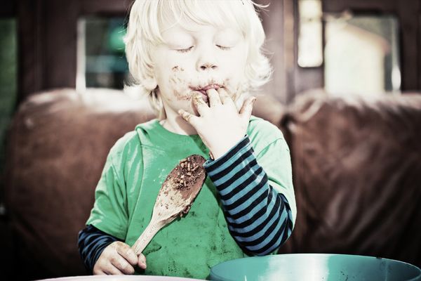 A small boy holding a cooking spoon in his hand, in a dirty shirt, licking his fingers