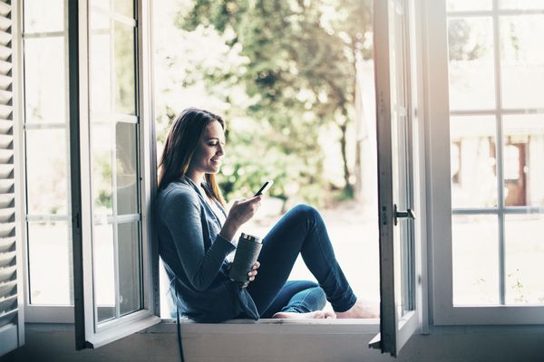 Uma mulher jovem sentada num lugar à janela, que se encontra aberta, a utilizar a aplicação Home Connect