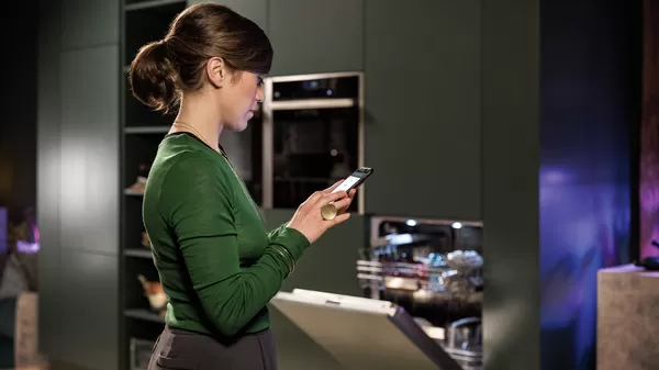 O persoană îmbrăcată într-un pulover verde ține un smartphone în mână, stând într-o bucătărie stilată cu electrocasnice moderne.