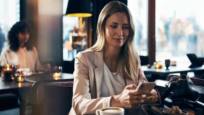 Una mujer sentada en una cafetería mirando su teléfono móvil. Lleva una chaqueta de color claro y tiene una taza de café delante. Al fondo, otra persona está sentada en una mesa.