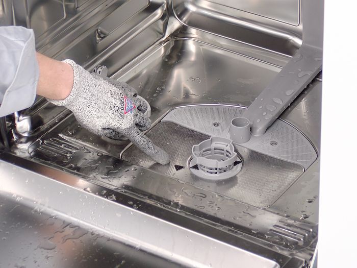 Mano con guanto che indica il filtro interno di una lavastoviglie, con gocce d&rsquo;acqua sulla superficie